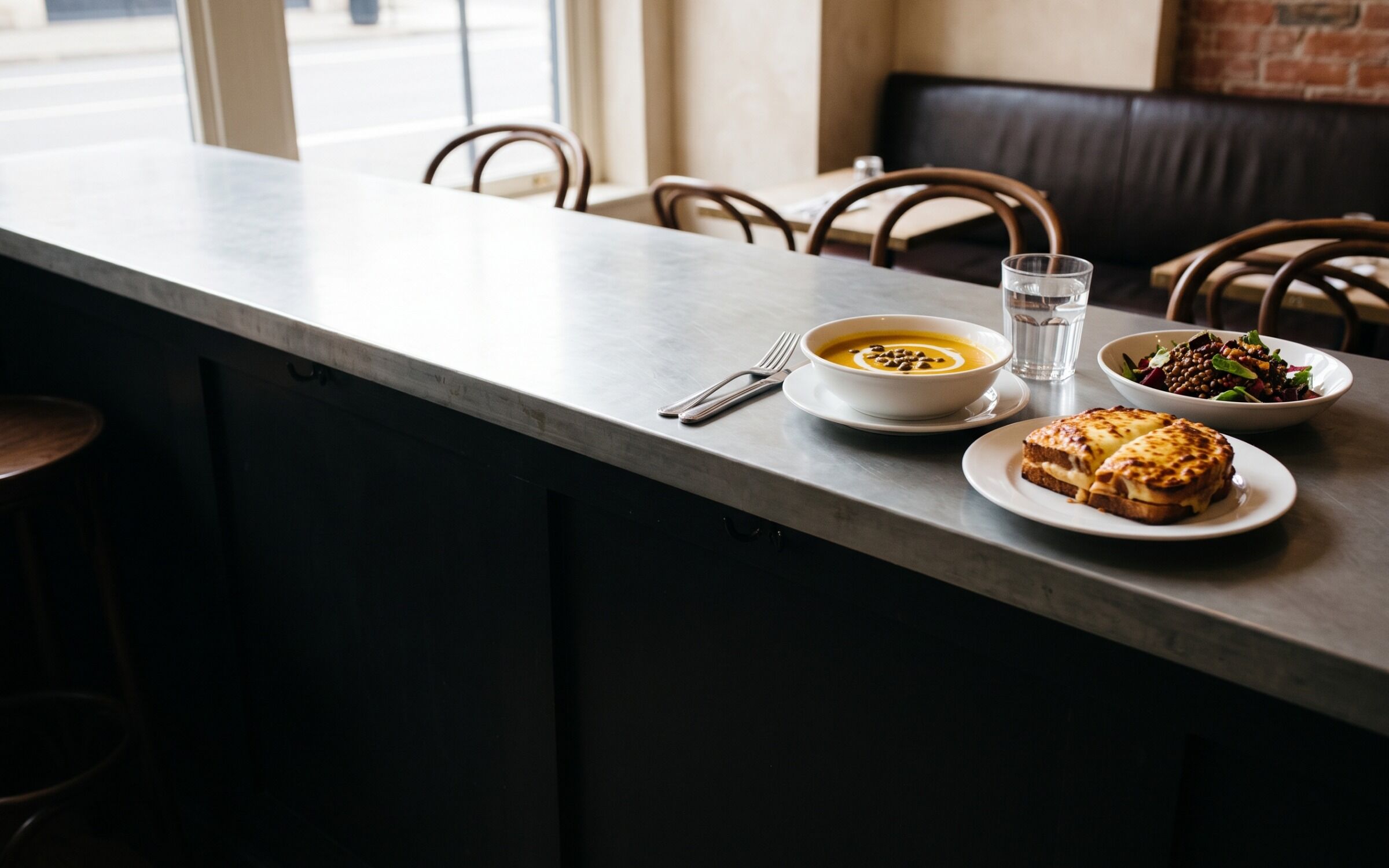 A bowl of soup, a croque cut in half and a lentil salad on the zinc, cutlery mid-use