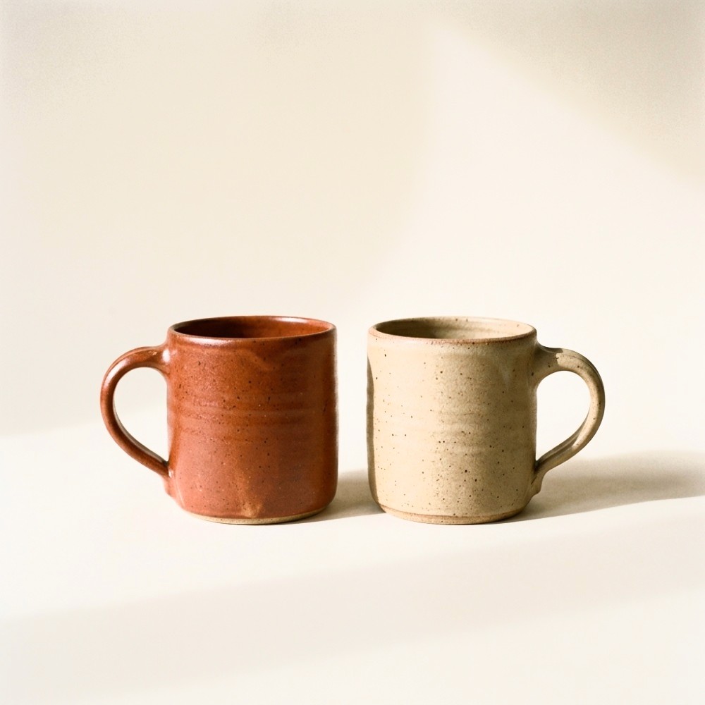 Two stoneware mugs with pulled handles standing side by side, one glazed terracotta and one sand.