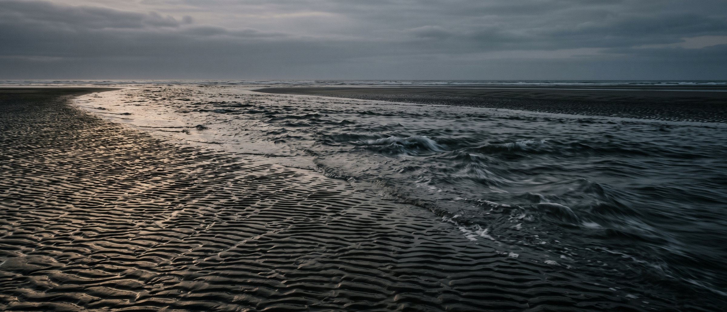 The incoming tide running fast up a wide sand channel at dusk, a sheet of water advancing over ribbed sand.