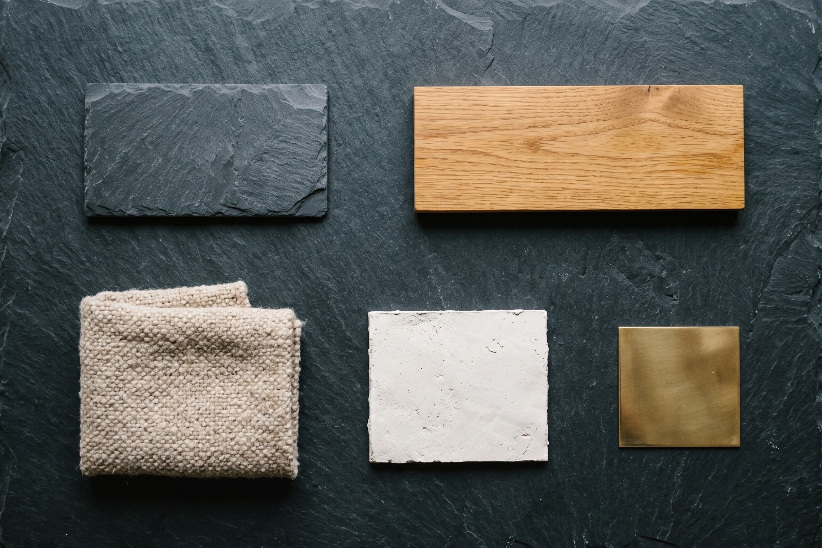 Five material samples laid out on dark slate and photographed from above: riven slate, oiled oak, folded oatmeal wool, a lime plaster tile, and a polished brass plate.